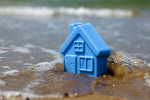 A blue toy house sits in the sand and is surrounded by water.